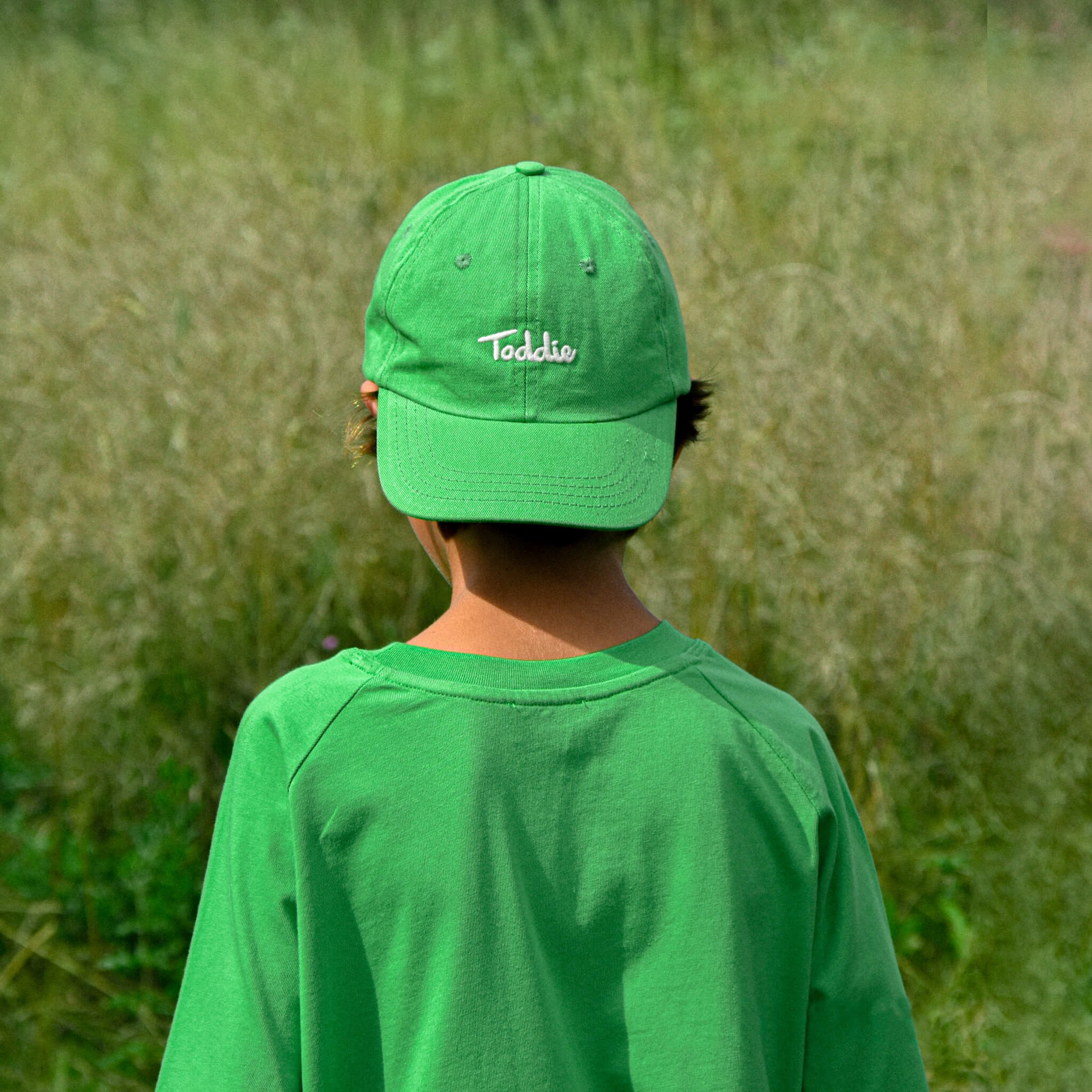 Child wearing vibrant green Toddie Cap with embroidered logo outdoors in a natural setting