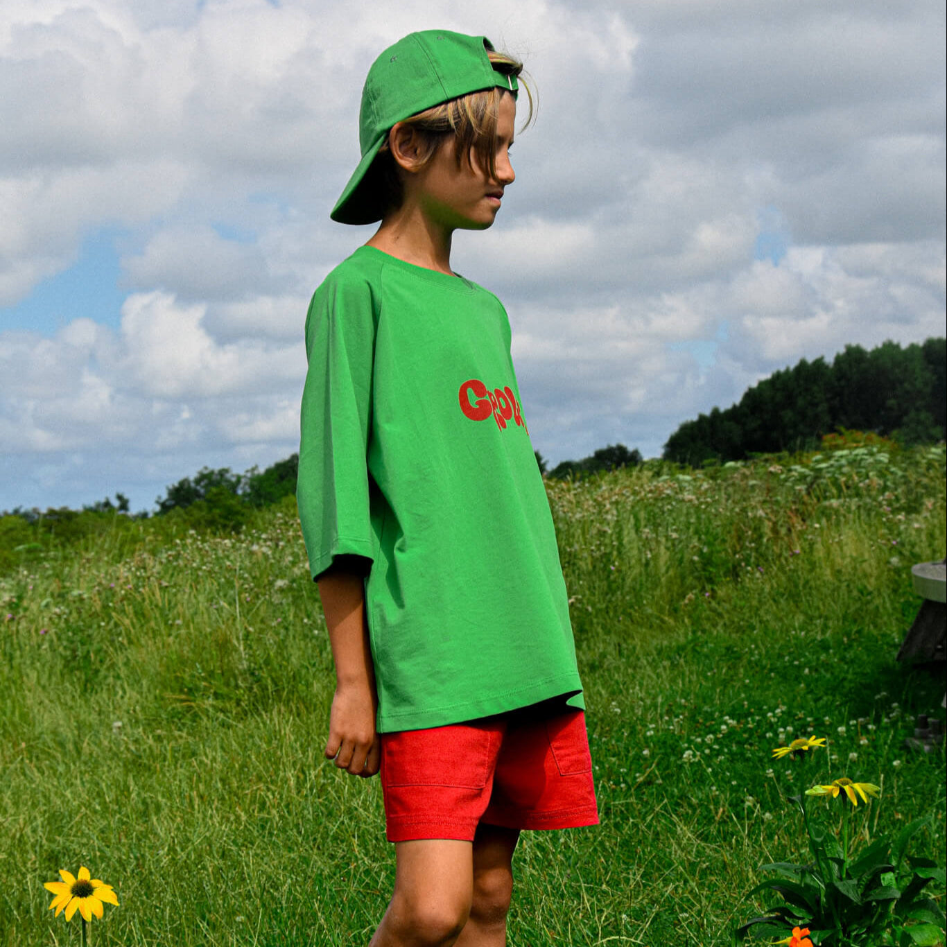 A child stands on grass under a cloudy sky, wearing a green cap backwards, an oversized green shirt with red text, and Toddie’s Baggy Short Red. Wildflowers and greenery surround them.