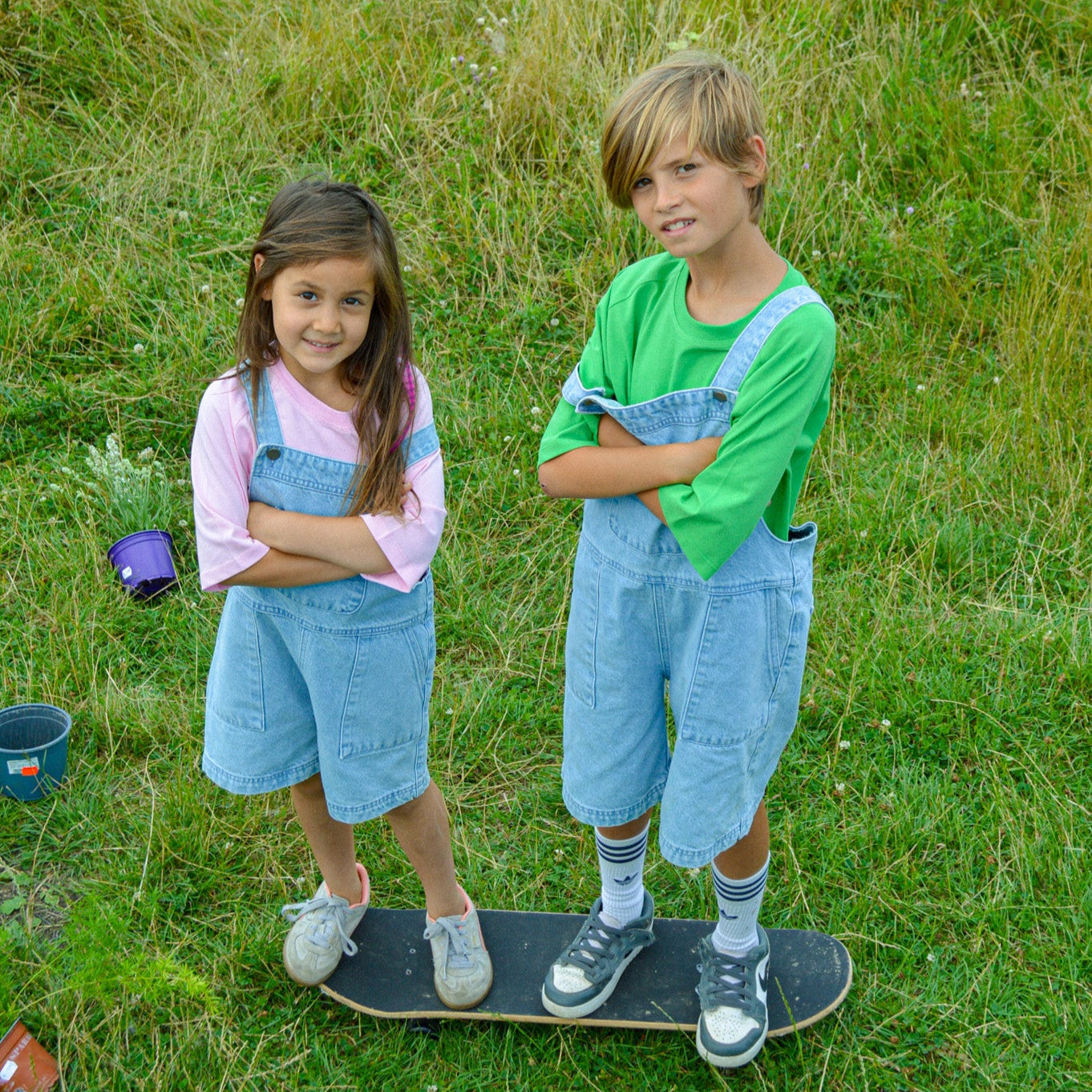 Two children stand on grass with arms crossed, wearing matching Toddie Denim Short Dungarees and colorful shirts. One stands on a skateboard near buckets, set against wild grass for a playful, comfortable look.