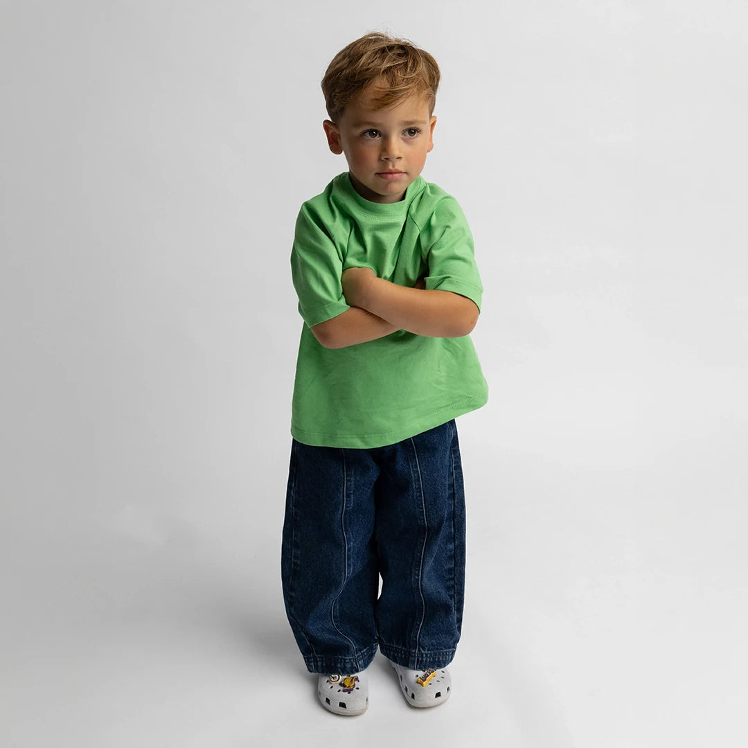 Young boy wearing Balloon styled - Denim Jeans with a green shirt and casual shoes on a plain background