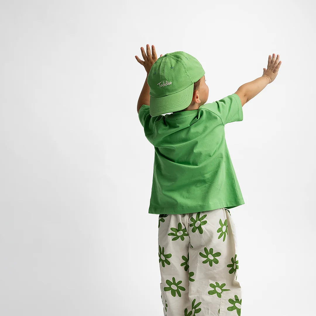 Child wearing a vibrant green Toddie Cap with embroidered logo and matching green shirt and floral pants.