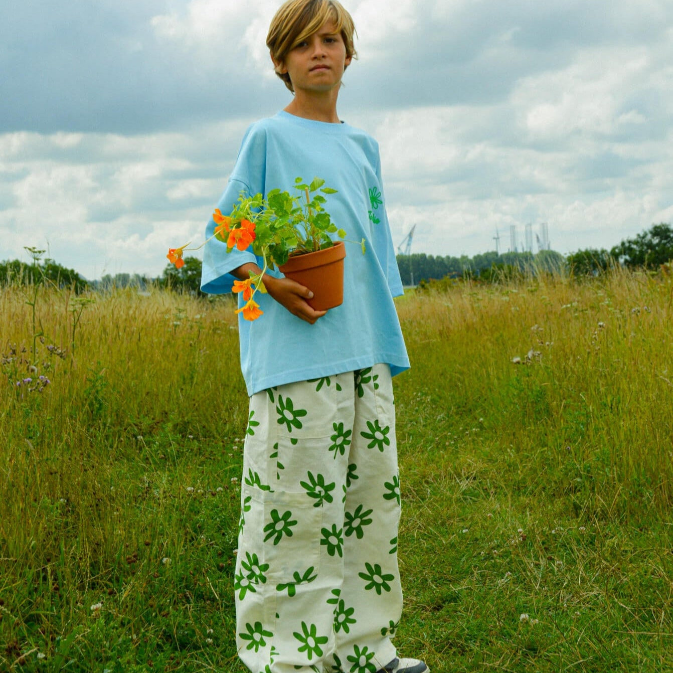Child wearing Cargo Flower Pants with green flower print holding a potted plant in a grassy field.