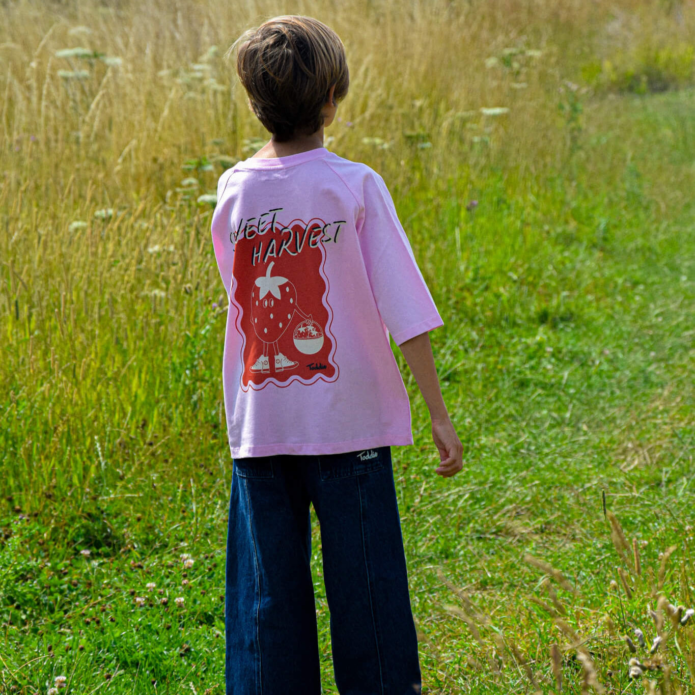Child wearing Sweet Harvest - Oversized Tee Pink with strawberry sneaker graphic in a grassy outdoor setting