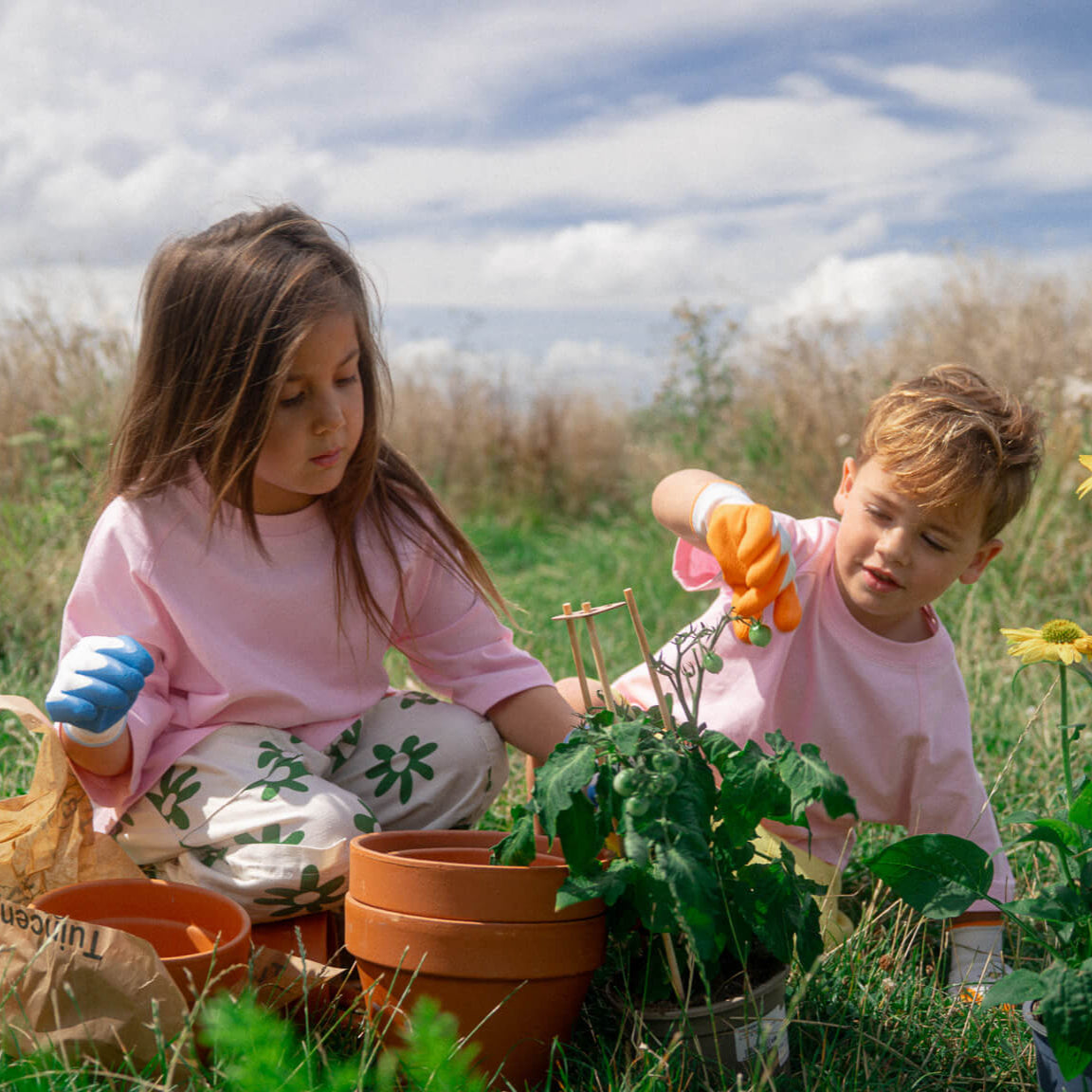 Two children wearing pink Oversized Tee gardening outdoors on a sunny day with plants and pots around them