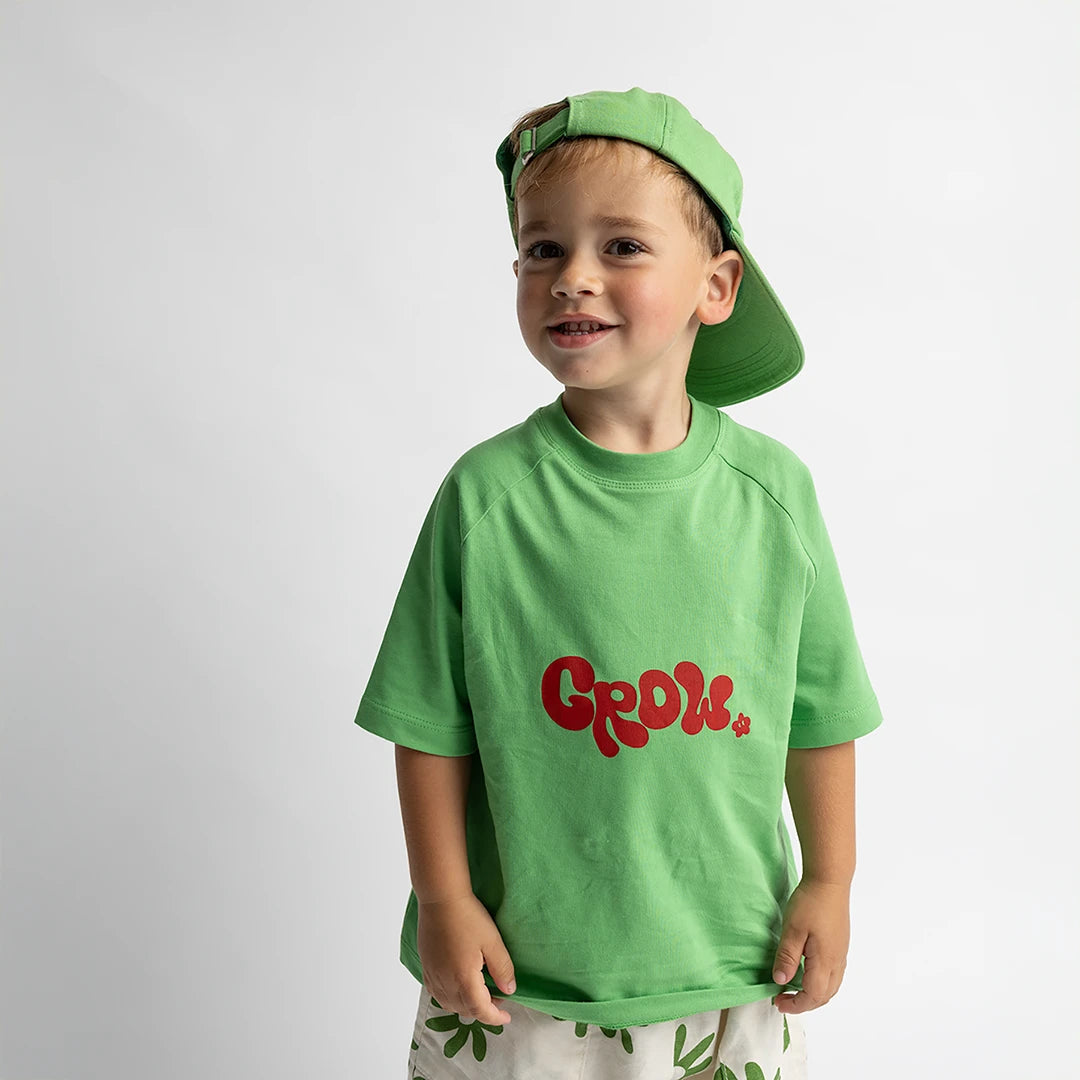 Child wearing green Grow - Oversized Tee with red lettering and matching green cap, smiling and standing against white background.