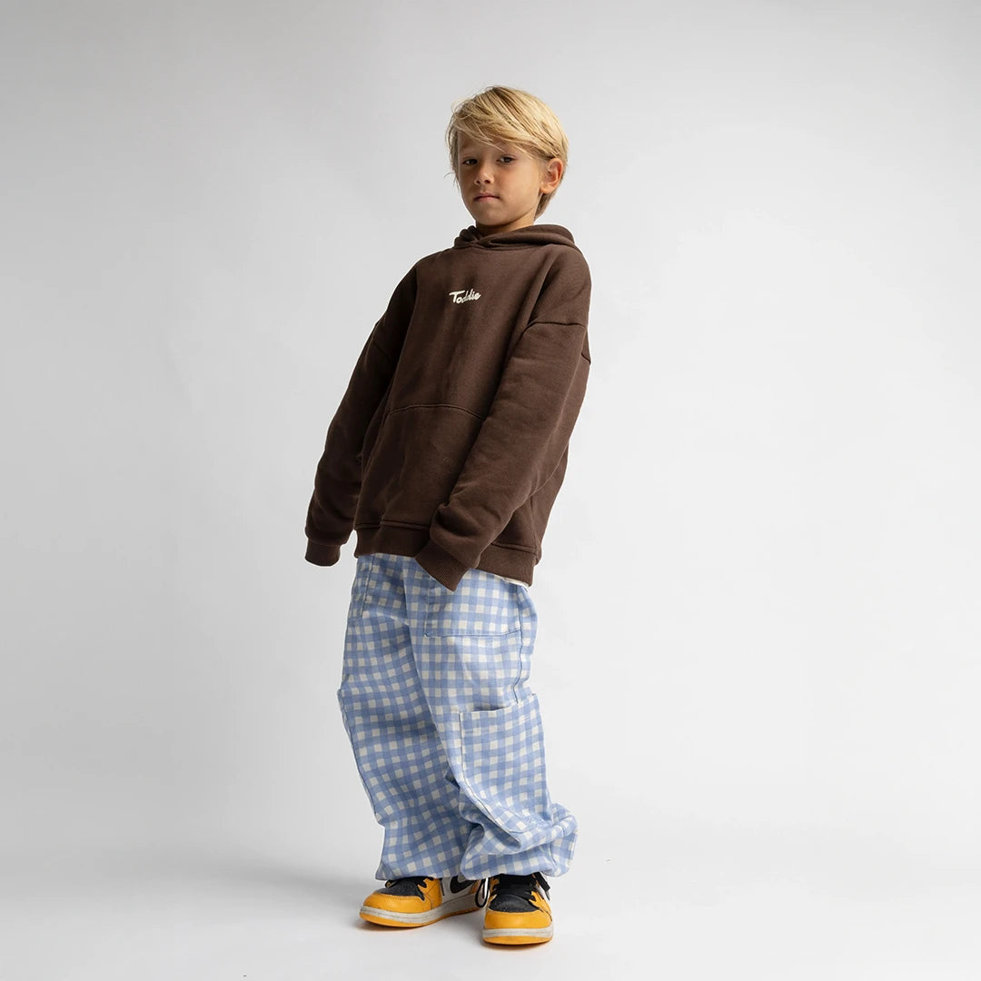 Child wearing a brown Toddie - Oversized Hoodie with logo, paired with blue checkered pants and yellow sneakers.