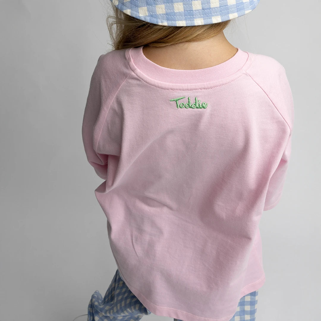 Child wearing a pink Oversized Tee with Toddie embroidery on back, paired with blue gingham pants and hat.