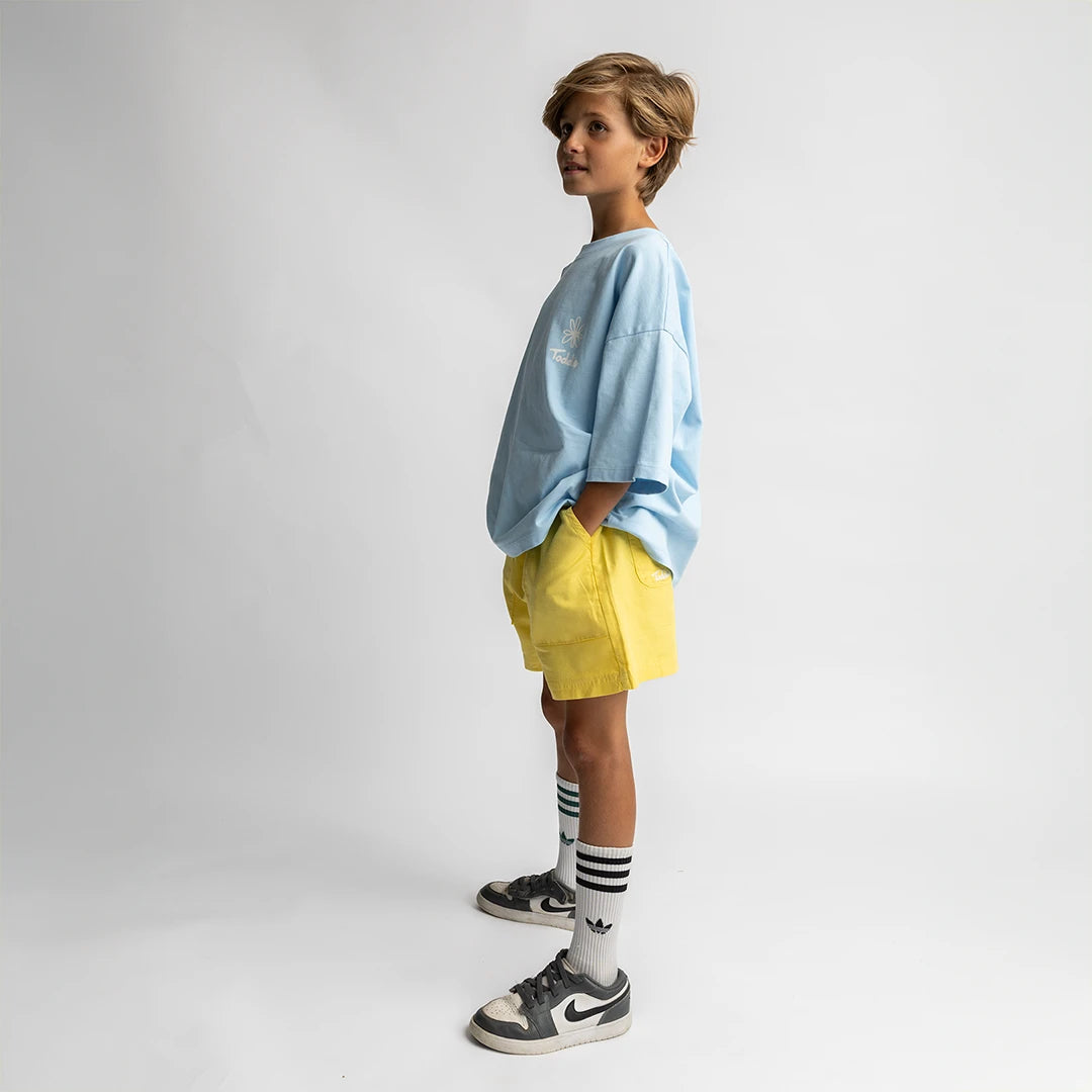 Side view of a kid wearing bright yellow baggy shorts with pockets, paired with casual sneakers and a loose blue tee Toddie Baggy Short Yellow